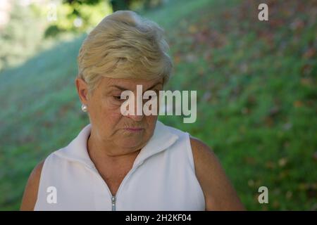 Sad senior woman looking down at park. Depressed elder lady wearing sleeveless white shirt. Pessimistic, worry, serious mother, blonde older person Stock Photo