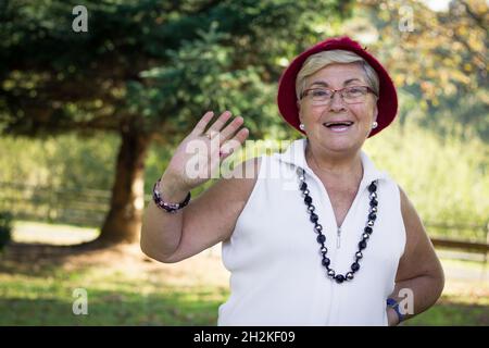 Senior smiling lady with red hat and eyeglasses waving on sunny day in the park. Older woman looking at camera with welcoming expression Stock Photo