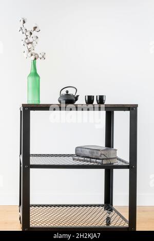 Industrial style trolley with green bottle with decorative cotton branch and black tea set with old books and white wall in the background Stock Photo