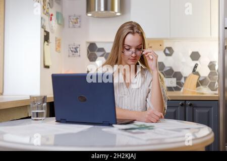 Young woman businesswoman signs documents while working from home in the kitchen with laptop Stock Photo