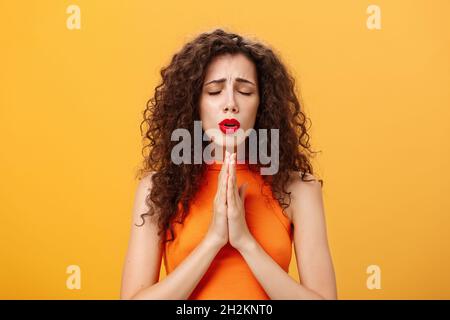 Nervous and concerned woman with curly hairstyle feeling hopeful praying with closed eyes and frowned eyebrows holding hands in pray near chest Stock Photo