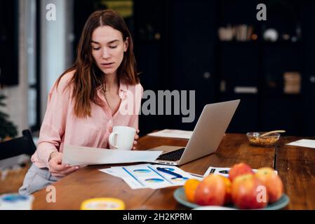 Woman working at home office sitting at desk analyzing business statistics holding diagrams and charts using laptop Stock Photo