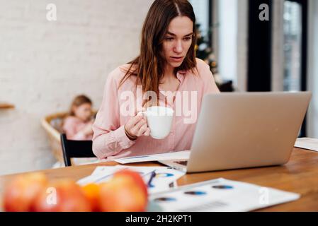 Young woman works in the kitchen on her laptop while her daughter stays home. Stock Photo
