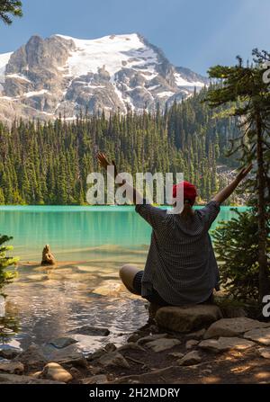 Happy young woman traveler outstretching arms feeling free, motivated looking at a beautiful landscape of the lake and mountains. Joffre Lake BC, Stock Photo