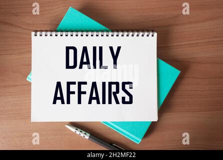 The text DAILY AFFAIRS is written on a notebook, next to it lies a notebook and a pen on a wooden table. Stock Photo