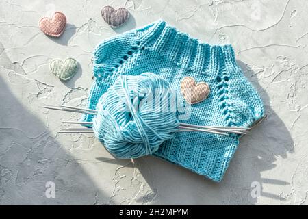 A knitted T-shirt for a toy made of turquoise cotton yarn. A ball of yarn, knitting needles and velvet hearts on a light green textured background Stock Photo