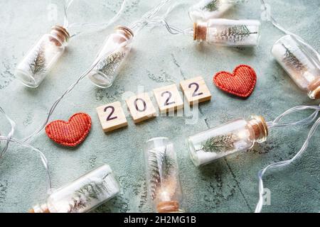 Number 2022 on wooden squares and a glowing garland of glass jars with Christmas trees and snow inside and two hearts on a textured green background Stock Photo