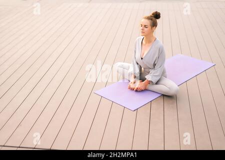 Wide shot of calm young woman sitting on yoga mat with closed eyes and meditating in butterfly pose, tilting head forward. Stock Photo