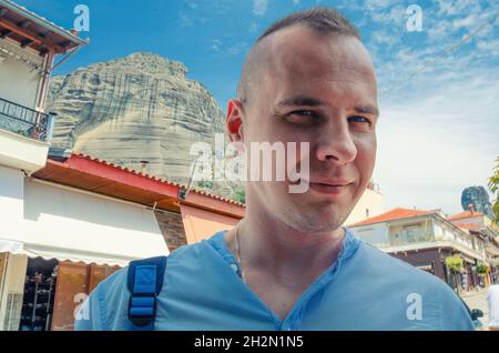 Close-up portrait of young man looking at camera, posing and smile outdoor in Kalabaka city, buildings, houses and Meteora stone rocks and mountains b Stock Photo