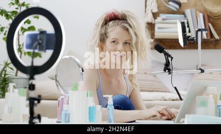 smiling woman influencer blogger recording a video tutorial on smartphone camera explains what to do to apply makeup cosmetics. Female vlogger making Stock Photo