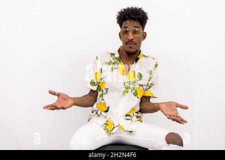 Young surprised Afro-American male in a floral shirt and sunglasses sitting with a white background Stock Photo