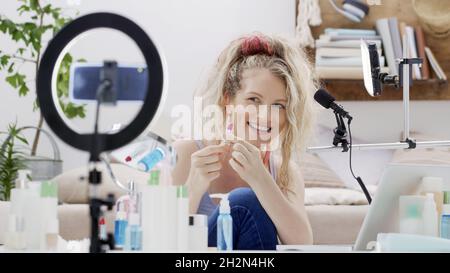 Blonde smiling woman influencer blogger recording a video tutorial on smartphone camera showing lipstick. Female vlogger making a beauty vlog for make Stock Photo