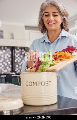 Mature Woman In Kitchen Making Compost Scraping Vegetable Leftovers Into Bin Stock Photo