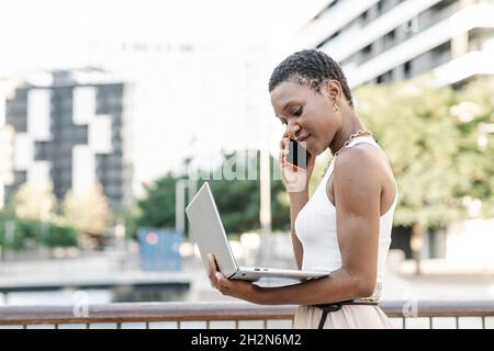 Female freelancer with laptop talking on mobile phone while standing by railing Stock Photo