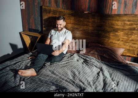 Smiling mid adult man using laptop while sitting on bed Stock Photo