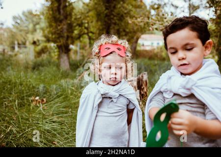 Blond boy wearing cape looking at male friend holding mask in forest Stock Photo