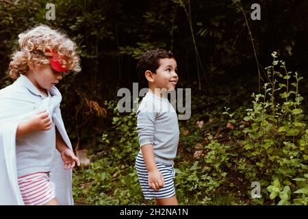 Blond boy wearing cape walking with male friend in forest Stock Photo