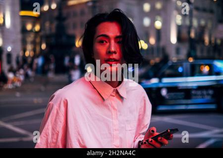 Handsome young man with long black hair holding mobile phone in city at night Stock Photo