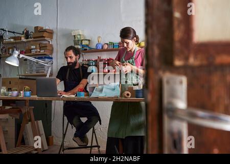 Female design professional packing box by male entrepreneur using laptop at workbench Stock Photo