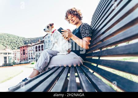 Mid adult woman sitting with pet dog while using smart phone on bench Stock Photo