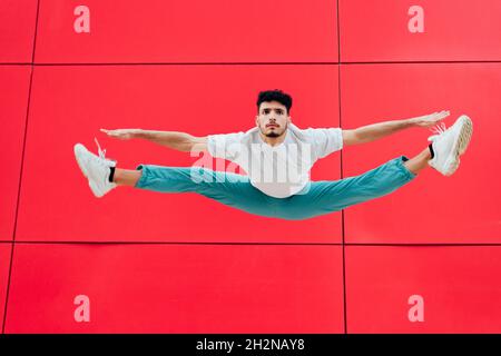 Man with arms outstretched doing splits in front of red wall Stock Photo