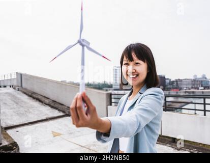 Smiling businesswoman holding wind turbine model on rooftop Stock Photo