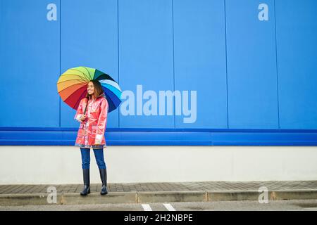 Woman with multi colored umbrella standing on footpath by blue wall Stock Photo