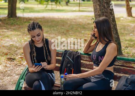 Hot attractive women smiling and swiping on their phones while sitting in a park and taking a break from their sports activities. Stock Photo