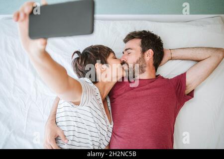 Affectionate couple kissing while taking selfie on bed at home Stock Photo