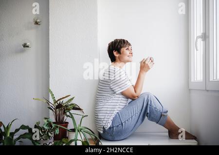 Smiling mid adult woman holding mug while sitting by plants at home Stock Photo