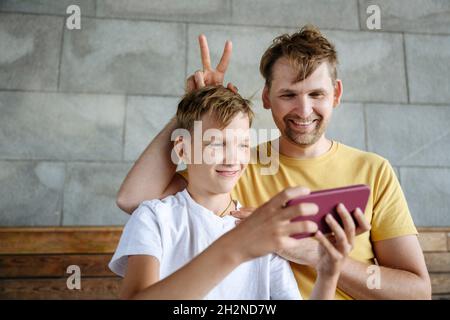 Smiling boy feeding iced coffee to father while sitting on bench Stock Photo