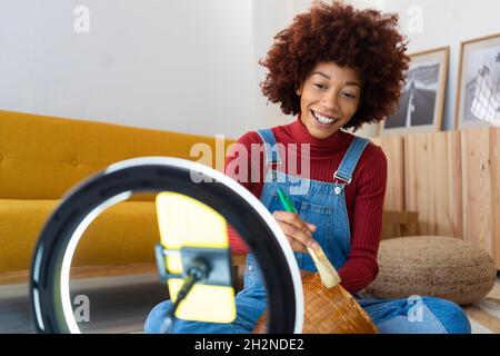 Female influencer vlogger painting wicker basket while live streaming through smart phone Stock Photo