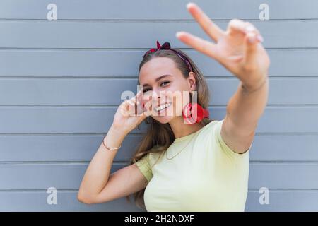Young woman gesturing peace sign in front of wall Stock Photo