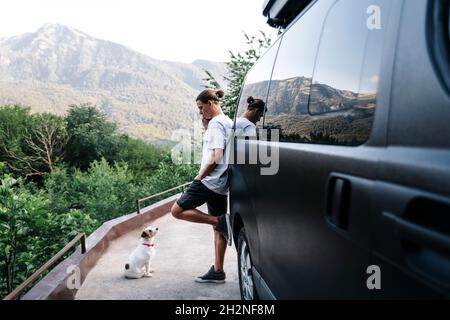 Man talking on smart phone looking at pet dog while leaning on car Stock Photo