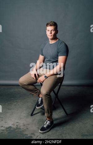 Young man sitting on chair in front of gray backdrop Stock Photo
