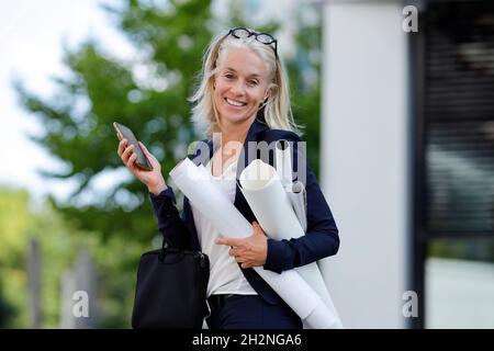 Smiling mature businesswoman carrying rolled up blueprints Stock Photo