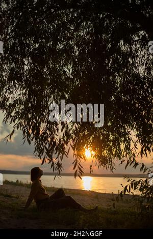 Woman sitting under tree during sunset Stock Photo