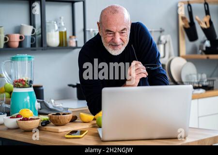 Cheerful senior man using laptop at kitchen counter Stock Photo