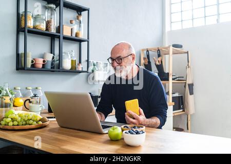 Happy senior man holding apple while using laptop at home Stock Photo