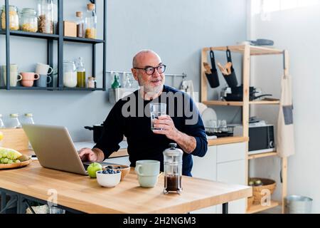 Smiling senior man using laptop at kitchen counter Stock Photo