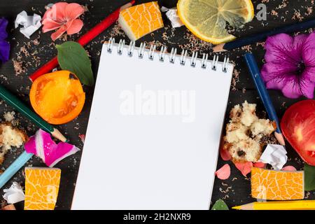 Paper notebook with blank pages, crumpled paper balls and crayons, scattered foods on wooden background, creative cooking recipe writing concept. Comi