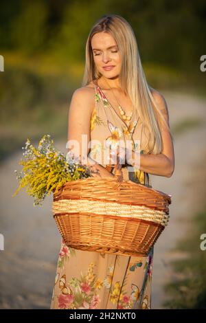 A young herbalist stands on a sandy road with a basket full of herbs. Goldenrod and winter glory. She is facing the camera.