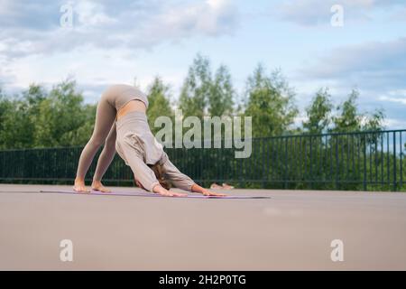 Flexible young woman sitting performing Downward-Facing Dog outdoors in city park standing on yoga mat. Stock Photo