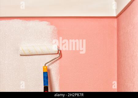 Close-up of an old fleecy paint roller painting pink wallpaper on the wall with white paint under the ceiling. Copy space. Stock Photo
