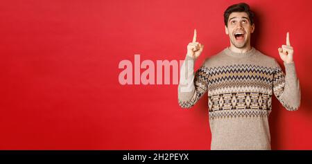 Image of excited handsome guy who likes winter holidays showing advertisement, looking and pointing fingers up with amazed face, standing in christmas Stock Photo