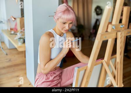Thoughtful woman with pink hair looks at canvas holding pencil in light studio Stock Photo
