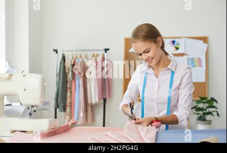 Portrait of young woman seamstress who uses scissors to cut out detail of dress on lines of sketch. Stock Photo