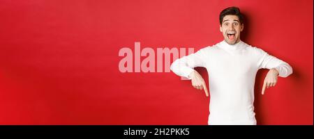 Image of amazed handsome guy in white sweater, reacting excited to christmas advertisement, pointing fingers down, showing logo, standing over red Stock Photo
