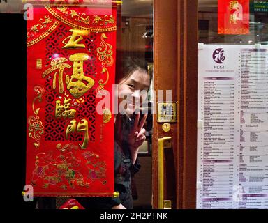 Chinese waitress at door of FLM Chinatown restaurant giving friendly welcome hello wave sign. Menu and prices in GBP on window to browse before entry. Chinatown Soho London UK Stock Photo