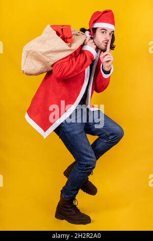 A young handsome man dressed up like Santa Claus with a sack full of presents invites to be silent and keep a secret by placing the index on his lips. Stock Photo